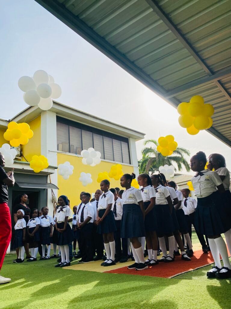 Pupils lined up at assembly in front of the yellow school building, decorated with yellow and white balloons