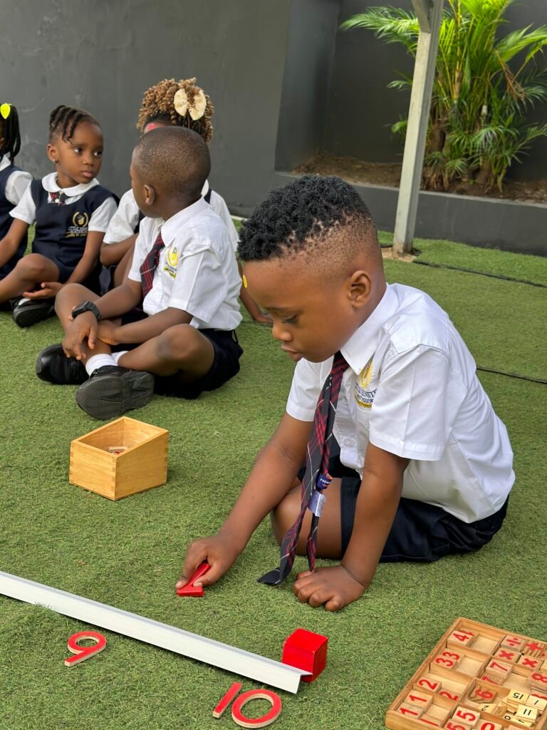 A pupil in uniform working with number rods and number cards on the lawn, with classmates behind