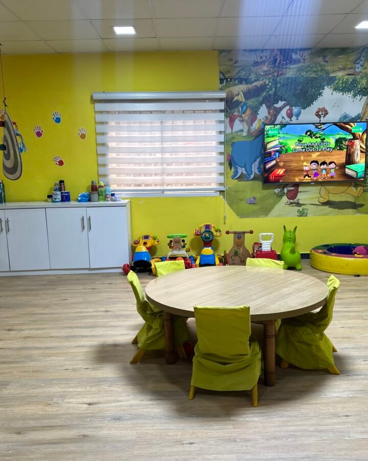 A bright early years room with a round table, small yellow chairs, soft play mats and a wall mural